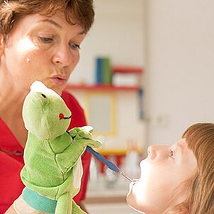 Zahnärztin führt bei einem kleinen Mädchen mithilfe eines Kuscheltier-Frosches Prophylaxe durch